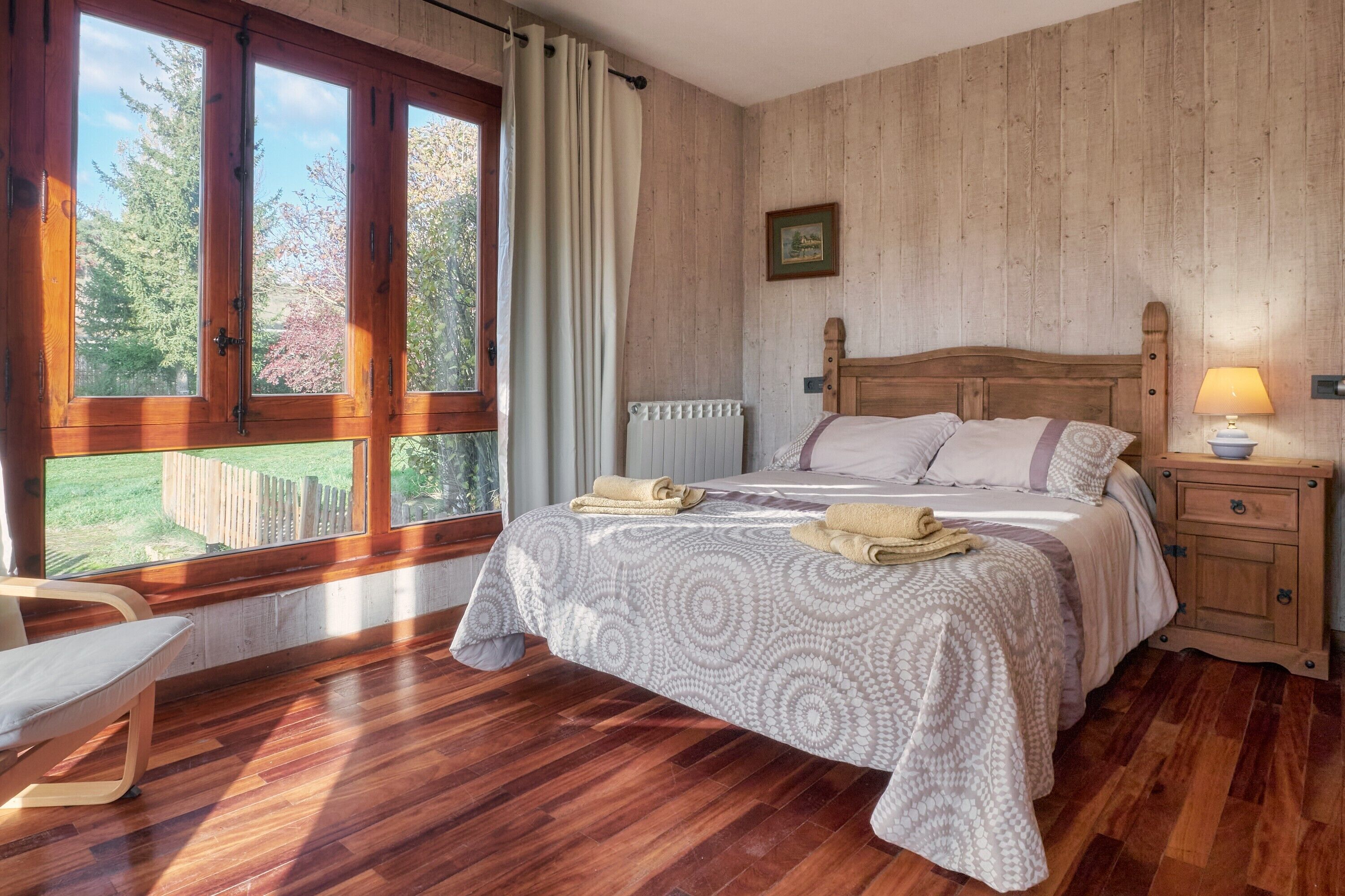 A bedroom with a wooden bed, a window with curtains, a bedside table with a lamp, and a chair.