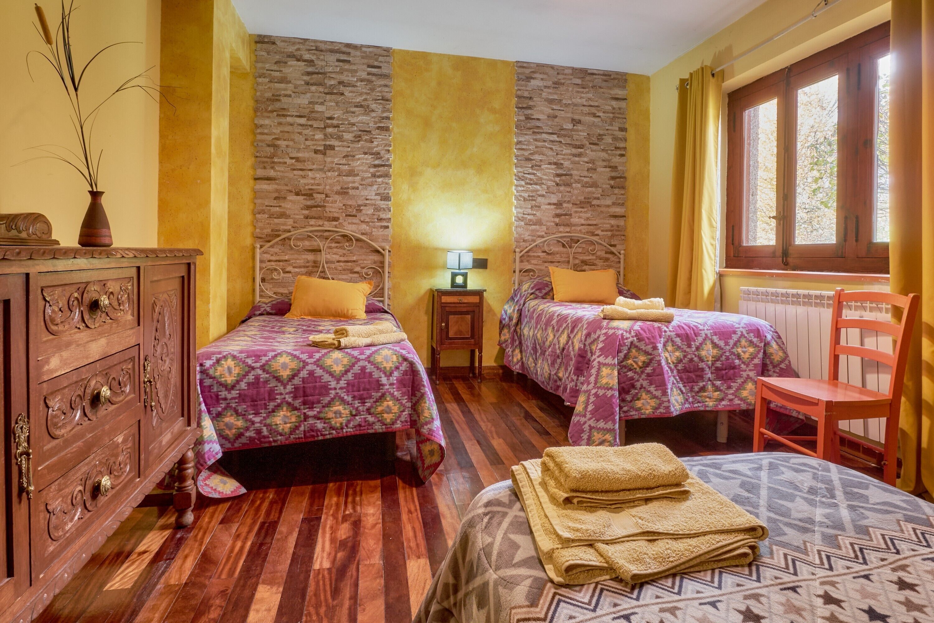 A room with two beds, a wooden dresser, a nightstand with a lamp, and a window with curtains.