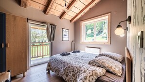 Una camera da letto con soffitto in legno, un letto con biancheria da letto floreale, una finestra vista su piante e un punto luce fissato al muro.