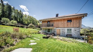 Una casa di legno a due piani con un balcone, immersa in un giardino curato e con le montagne sullo sfondo.