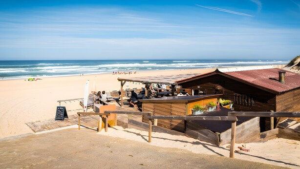 Una estructura de madera junto a la playa con un área de estar cubierta, un cartel de pizarra y vista al océano.
