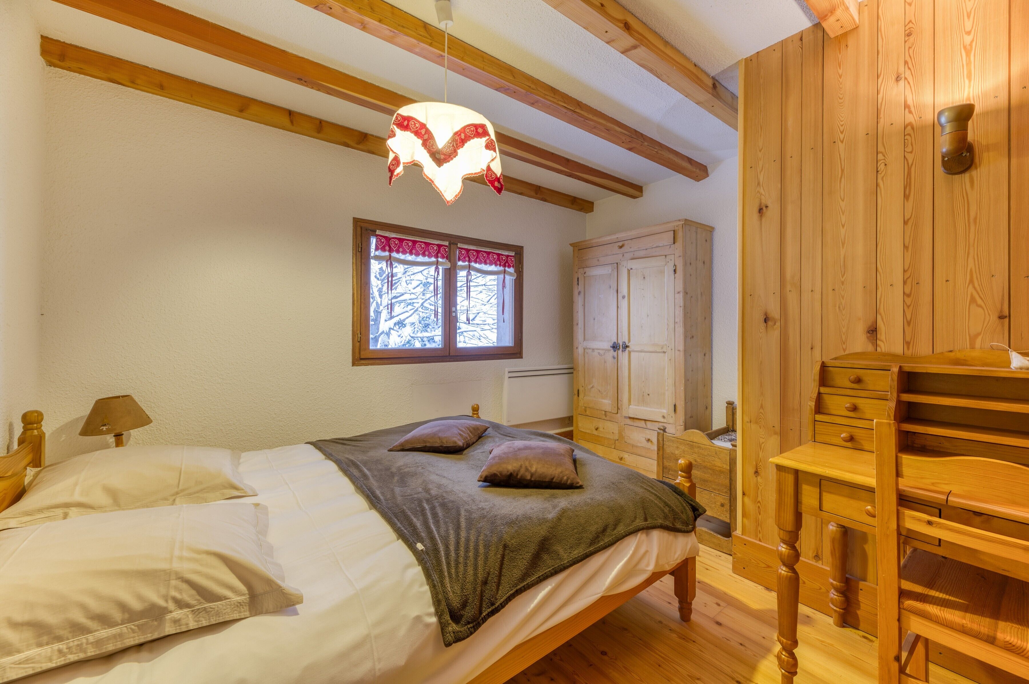 A bedroom with a bed, wooden headboard, a window with curtains, a wardrobe, and a desk with a chair.