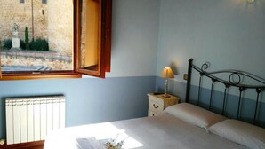 A bedroom with a bed, a nightstand, a lamp, a window with an open shutter, and a view of a building outside.
