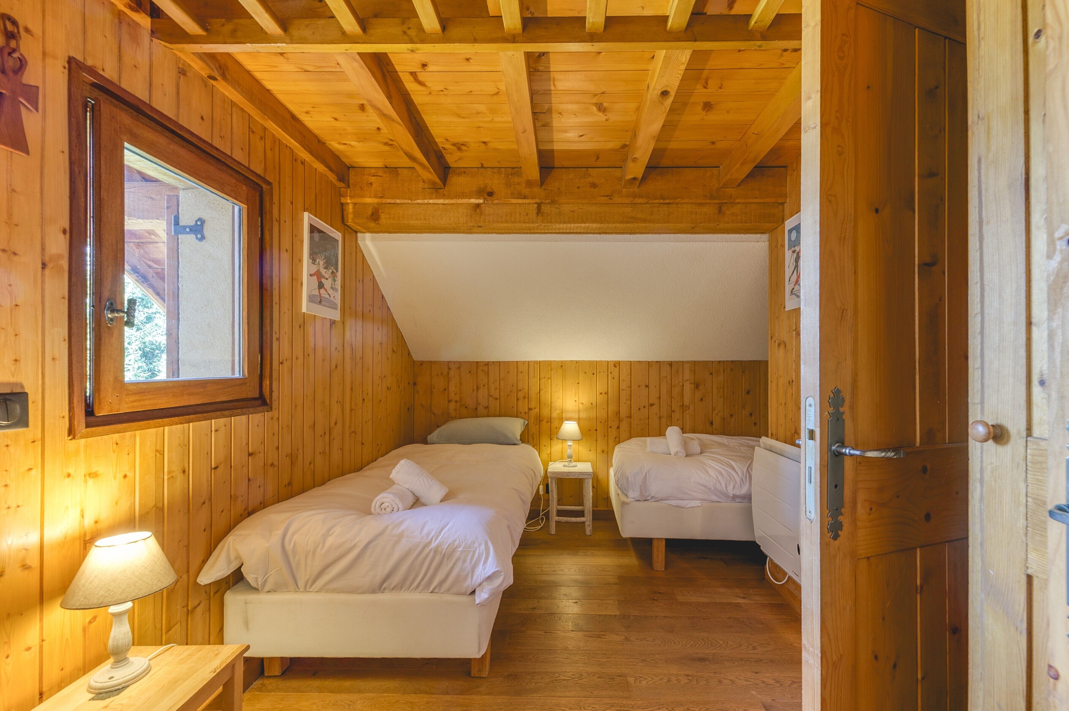 A wooden cabin room with two beds, a small table with a lamp, and a door.