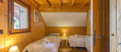 A wooden cabin room with two beds, a small table with a lamp, and a door.