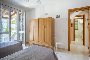 A bedroom with two beds, a wooden wardrobe, a white chair, and a bathroom visible through an open door.