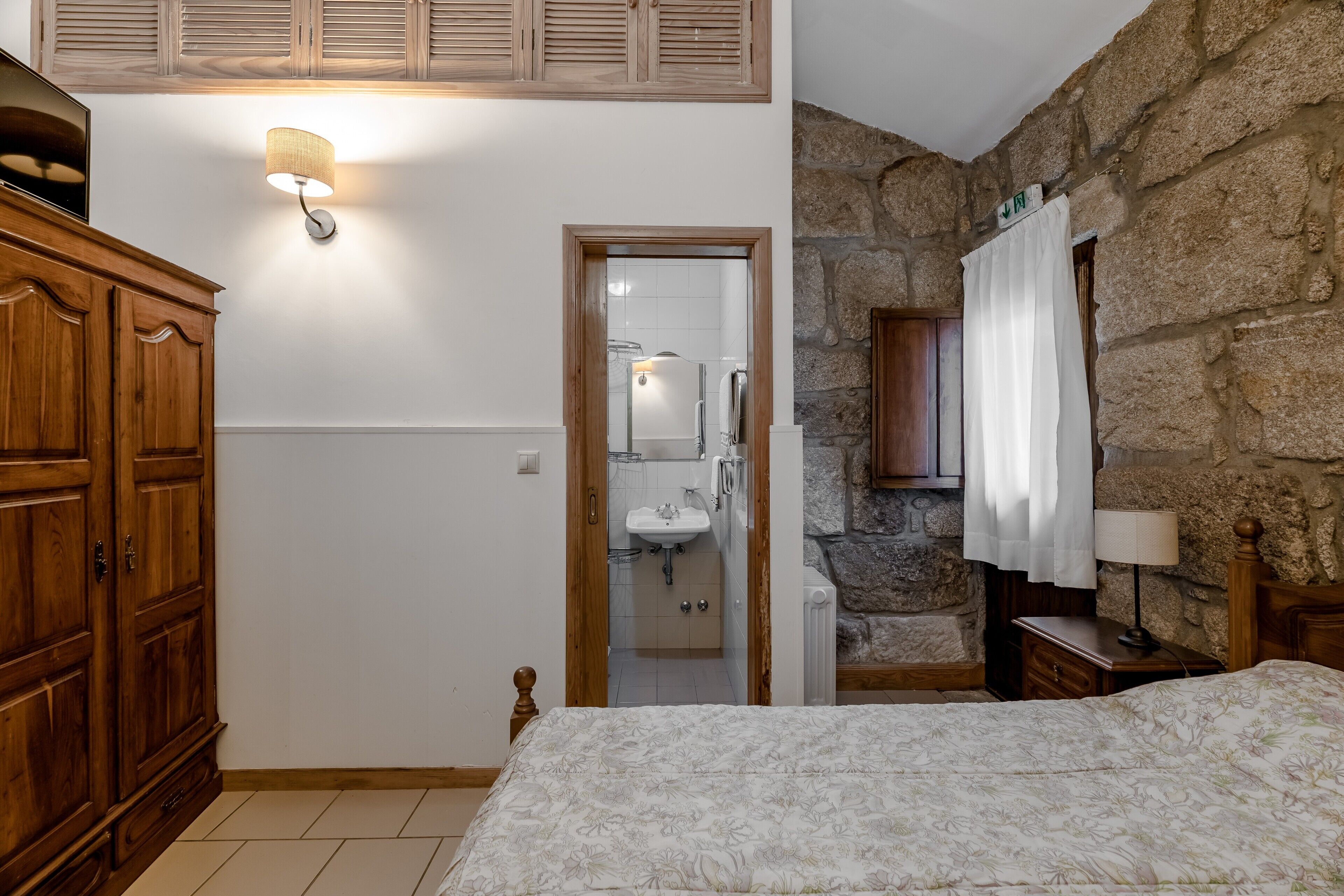 Un dormitorio con un armario de madera, una cama con cubrecama floral, una pared de piedra, una mesita de noche de madera y una lámpara de noche.