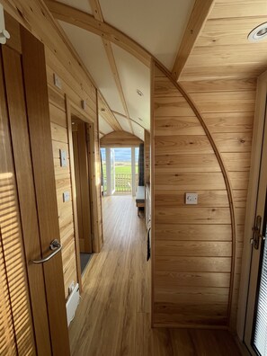 Interior de madera con techo alto, pasillo que lleva a un balcón y vista de un paisaje verde.