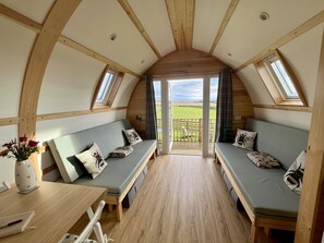Um quarto de loft acolhedor e semiaberto, com vigas de madeira, um sofá-cama, uma mesinha e vista para um campo verde através de uma porta de vidro.