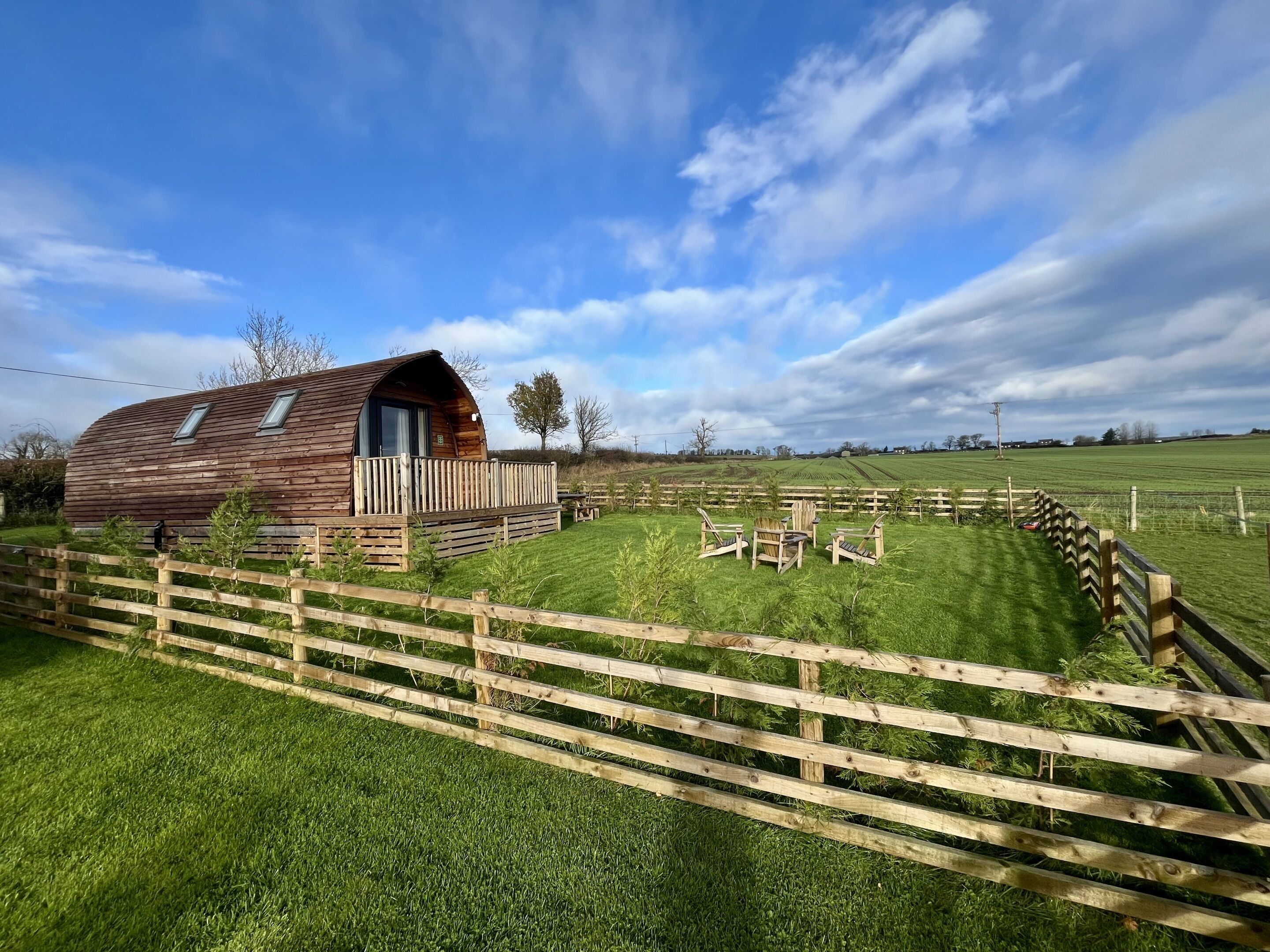 A wooden cabin with a sloped roof and a balcony, surrounded by a wooden fence and a grassy field.
