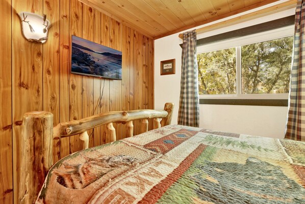 A wooden cabin room with a bed, a painting, a window with curtains, and a wall-mounted light.