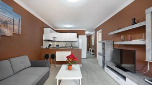 A modern living room with a grey sofa, a white coffee table with a potted plant, a TV on a built-in shelf, and a kitchen in the background.