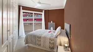 A bedroom with a bed, a ceiling fan, a bedside table with a lamp, and a view of a balcony with a red railing.