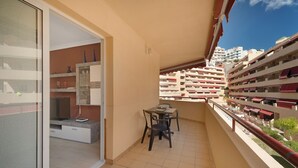 A balcony with a table and chairs, a glass door leading to a room, and a view of a multi-story building with balconies.