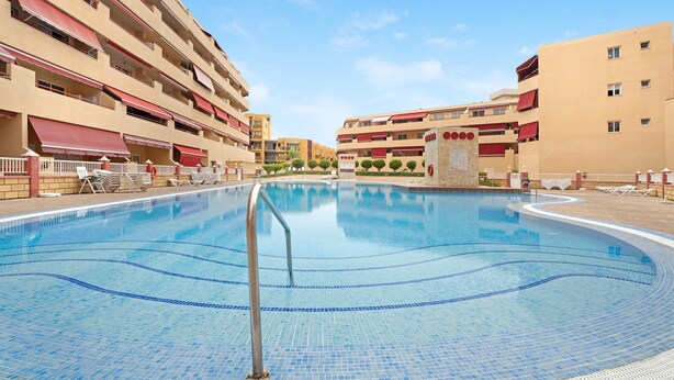 A swimming pool with a metal ladder, surrounded by beige buildings with red awnings and balconies.