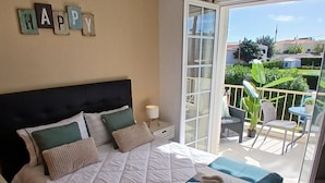 Een slaapkamer met een bed, kussens, een nachtkastje, een raam en een balkon met een stoel en een tafel.