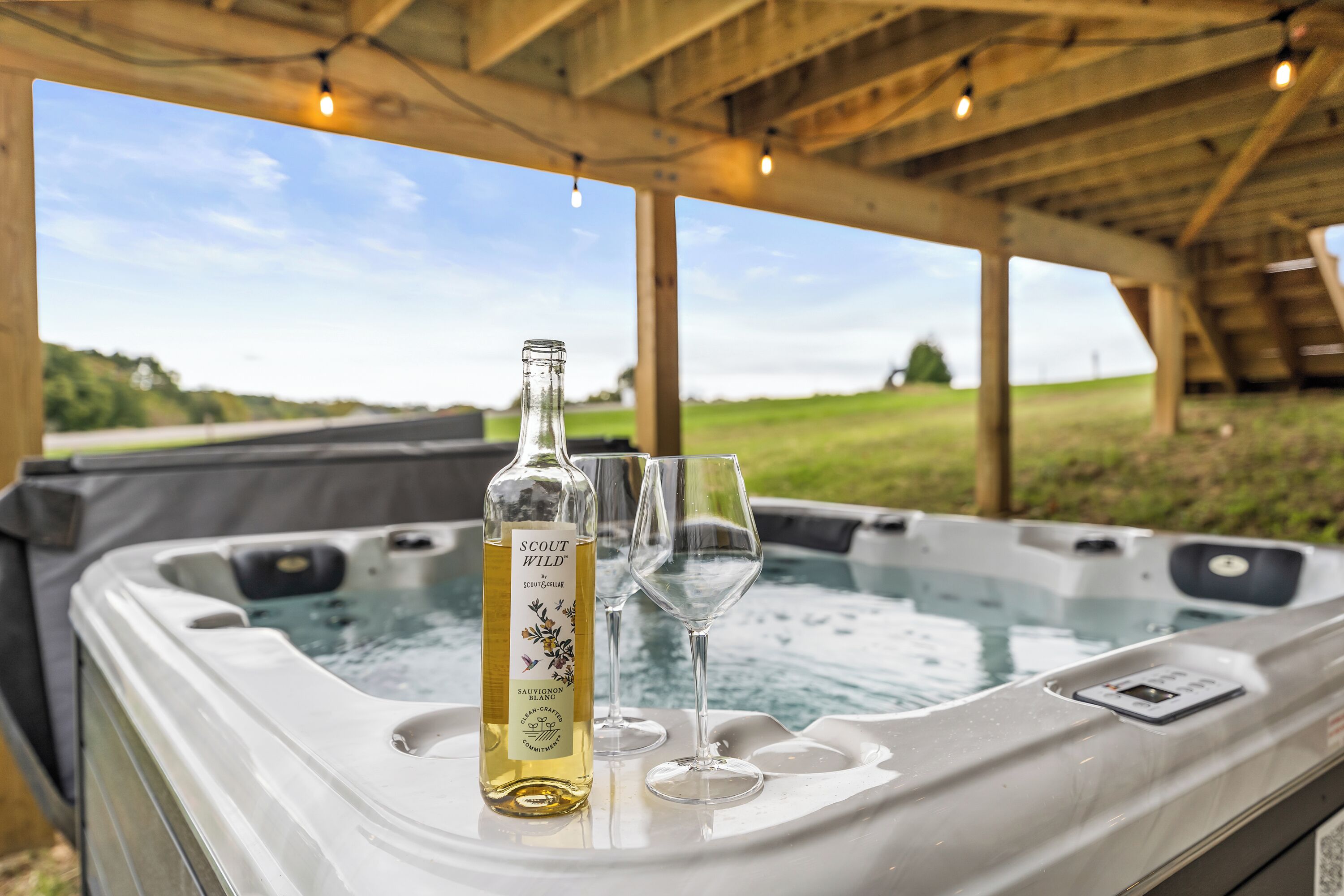 A hot tub with a bottle of 'Scout Wild' wine and two glasses, set outdoors with a view of a grassy field and a clear sky.