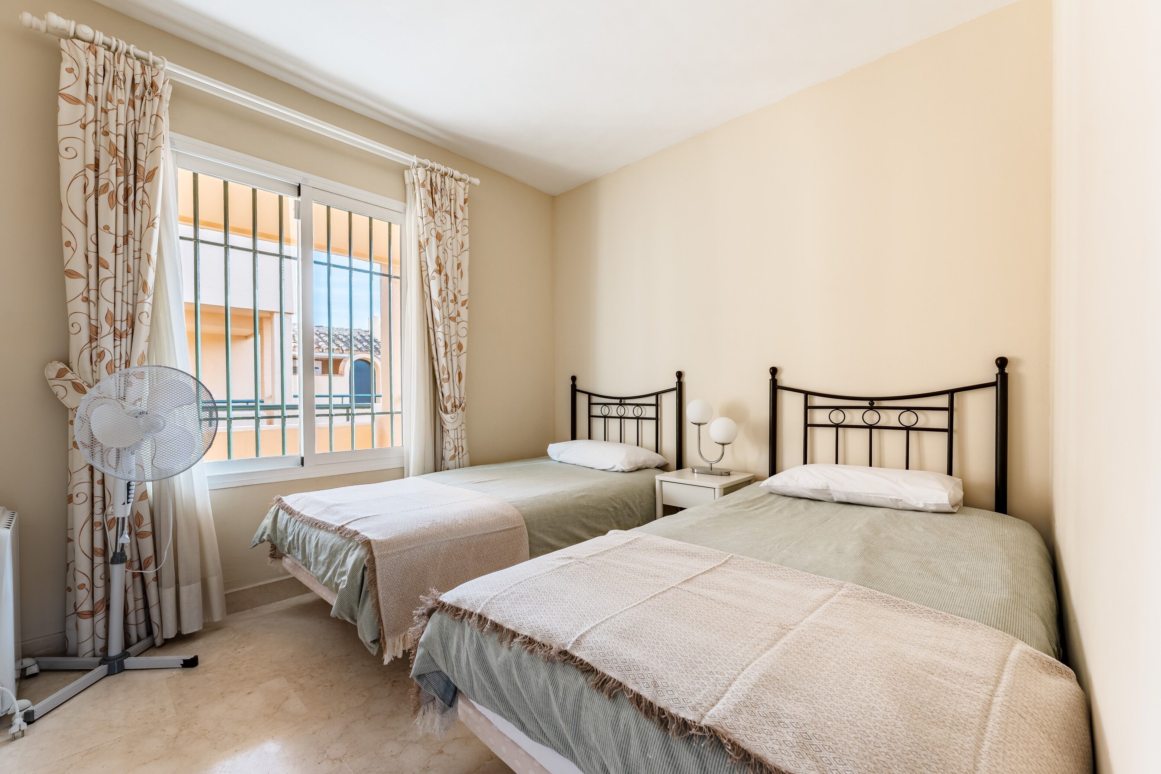 Een slaapkamer met twee bedden, een raam met gordijnen, een ventilator en een nachtkastje met een lamp.