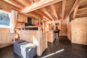 En stuga med träpanel på insidan, utrustad med ett litet kök, en bardisk, en sittgrupp med en bänk och ett fönster med utsikt över ett snötäckt landskap.