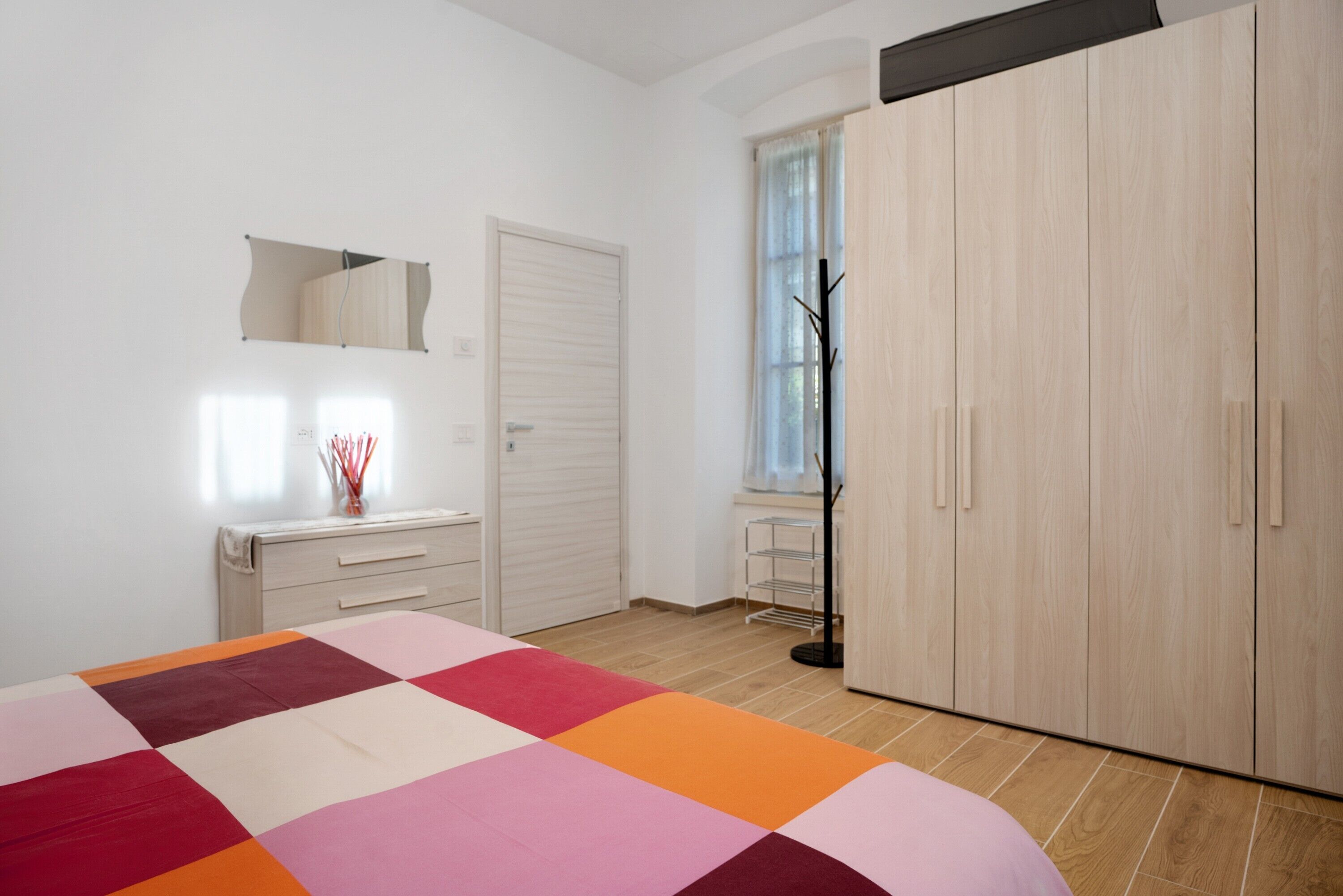 Un dormitorio con cama, armario, cómoda, espejo y ventana con cortinas.