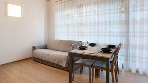 A room with a wooden floor, a beige sofa, a dining table with chairs, and sheer curtains. - Apartment 'Appartamento Chiara Tione' with Private Garden and Wi-Fi (Tione di Trento)
