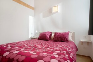 Een slaapkamer met een bed, kussens, een nachtkastje, een klein raam en een wandplank.