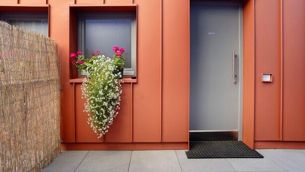 A building entrance with a metal door, a small window with flowers, and a bamboo fence. - Apartment 'Appartamento Chiara Tione' with Private Garden and Wi-Fi (Tione di Trento)