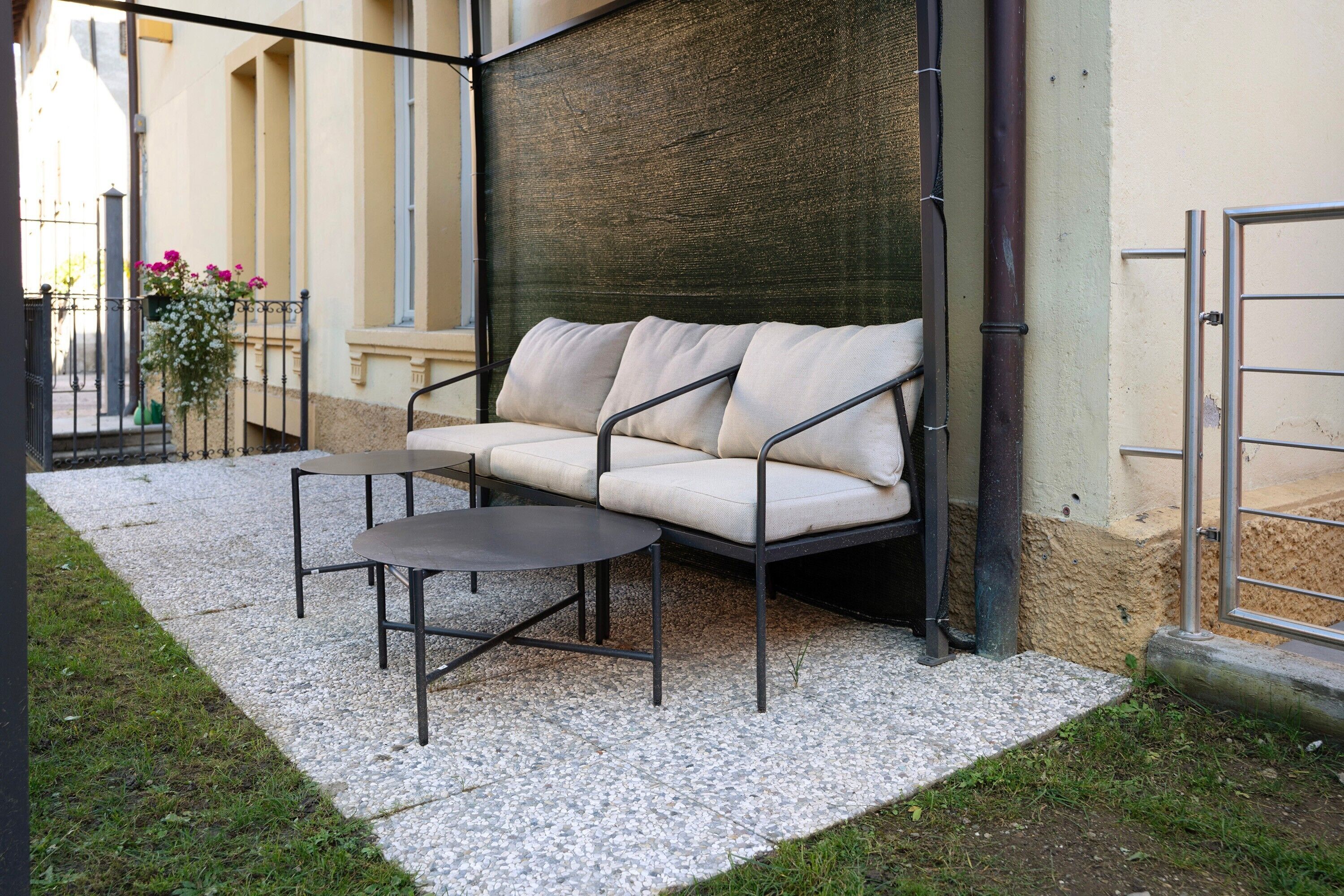 Un espace extérieur aménagé avec un canapé et une table, un sol en gravier et un bâtiment doté d’un mur à texture et d’un portail en métal.