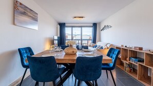 Een eetgedeelte met een houten tafel, blauwe stoelen, een vaas met bloemen en uitzicht buiten door grote ramen.