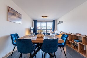 A dining area with a wooden table, blue chairs, a vase with flowers, and a view of the outside through large windows.