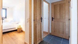 Een slaapkamer met een bed, nachtkastje en een raam. Houten deuren leiden naar een andere kamer.