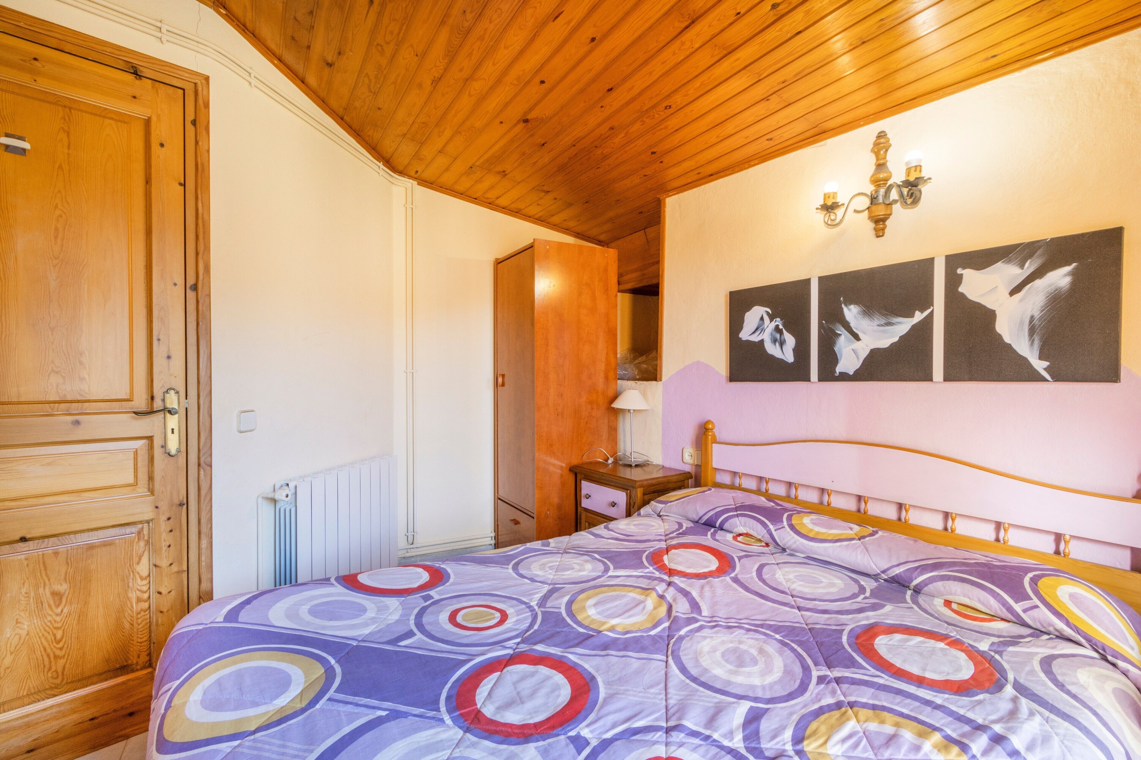 Un dormitori amb sostre de fusta, un llit amb col·locador de colors i un dibuix circular, una taula de nit amb una làmpada, i tres quadres penjats a la paret amb ocells.