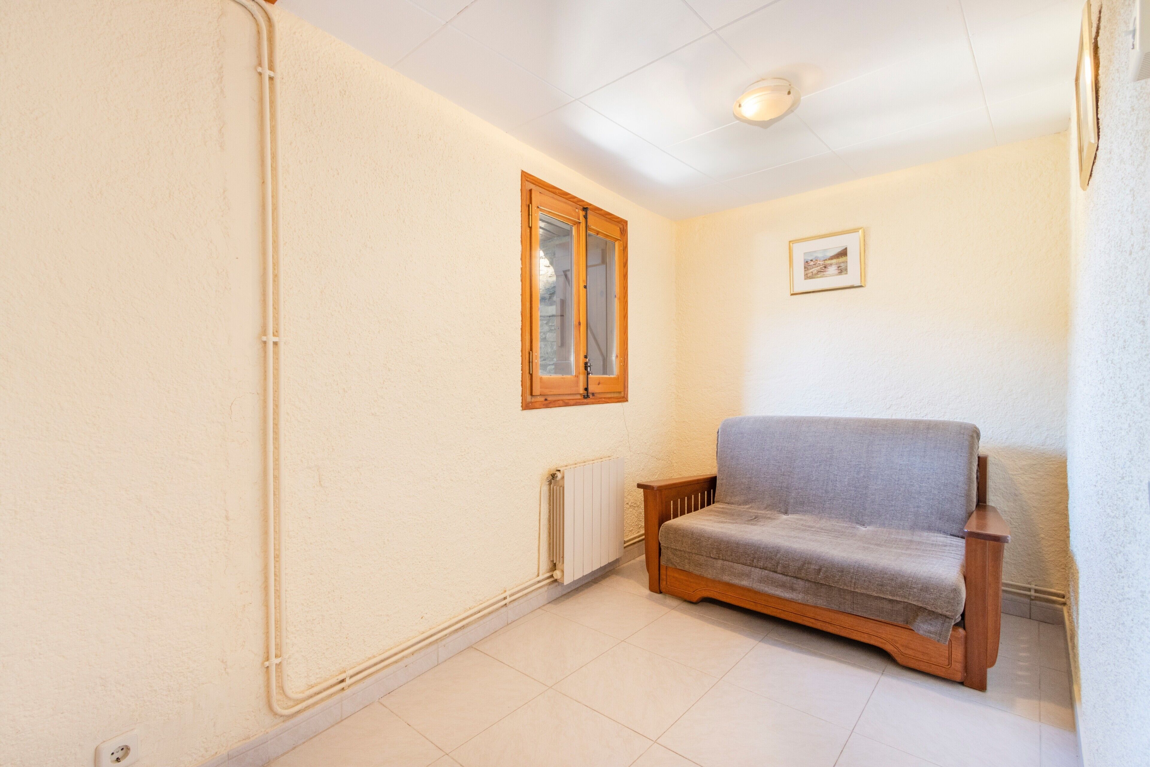 Une pièce avec une fenêtre à cadre en bois, un radiateur, un canapé gris et un tableau encadré accroché au mur.
