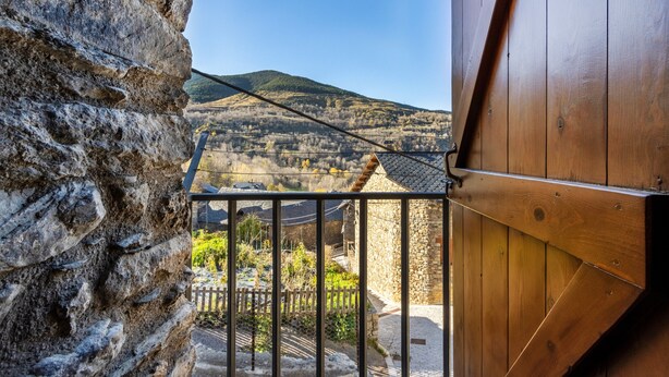 Un muro de piedra, barandilla de balcón de madera y vista a un paisaje montañoso.