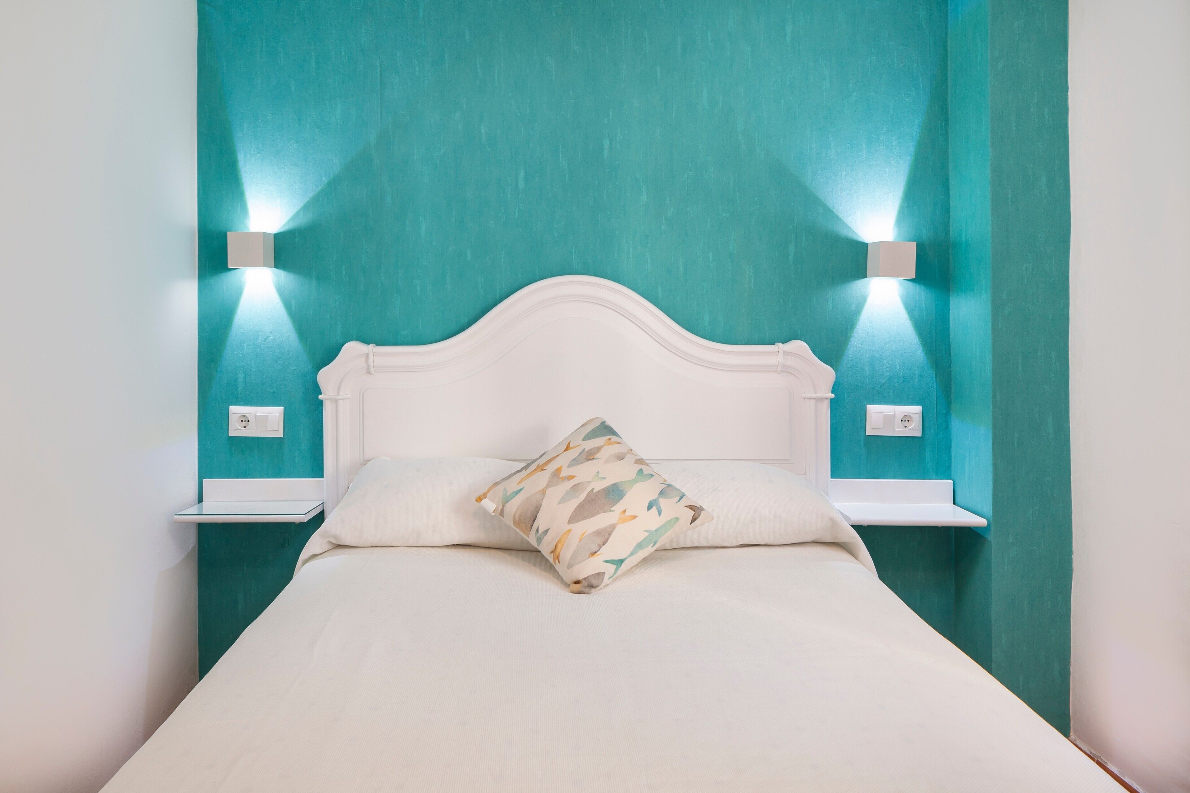 Una cama con cabecera blanca, almohada estampada y dos lámparas fijadas a paredes color turquesa.