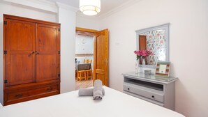 Une chambre avec une grande armoire en bois, un lit avec une serviette roulée, une coiffeuse avec un miroir, et un coin repas à l’arrière-plan.