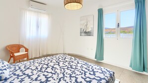 Un dormitorio con cama, una silla de mimbre, una ventana con cortinas, un equipo de aire acondicionado y un aplique de pared.