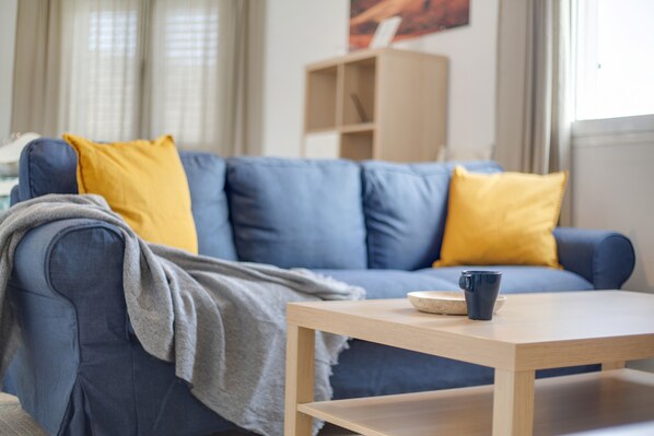 A living room with a blue sofa, yellow cushions, a wooden coffee table, and a bookshelf in the background.