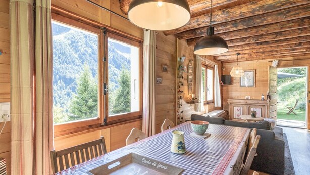 A wooden cabin interior with a large dining table, chairs, a kitchen area, and a view of a forested mountain. - Chalet "Le Petit Ranch" with Mountain View and Private Garden (Le Reposoir)