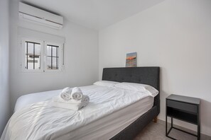 Un lit bien fait, avec du linge de lit blanc et une serviette enroulée, une tête de lit rembourrée de couleur grise, une petite table de chevet noire et un climatiseur fixé au mur.
