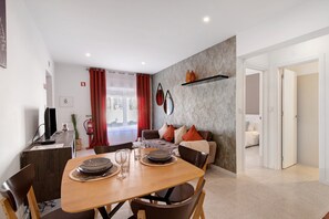 A modern dining area with a wooden table set for two, a sofa with cushions, a wall-mounted shelf, and a visible fire extinguisher.