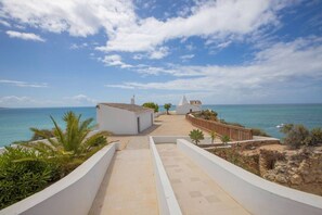 A coastal pathway leading to a white building with a conical roof, surrounded by greenery and overlooking the sea.