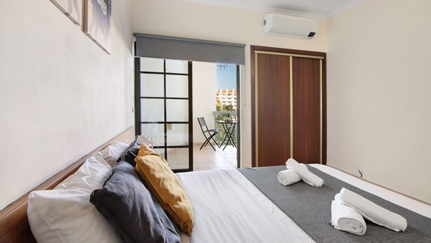 A hotel room with a bed, a small table, a chair, a sliding door to a balcony, and a wardrobe. - Sousa E Filhos Apartment – Shared Pool, Near the Beach (Albufeira)