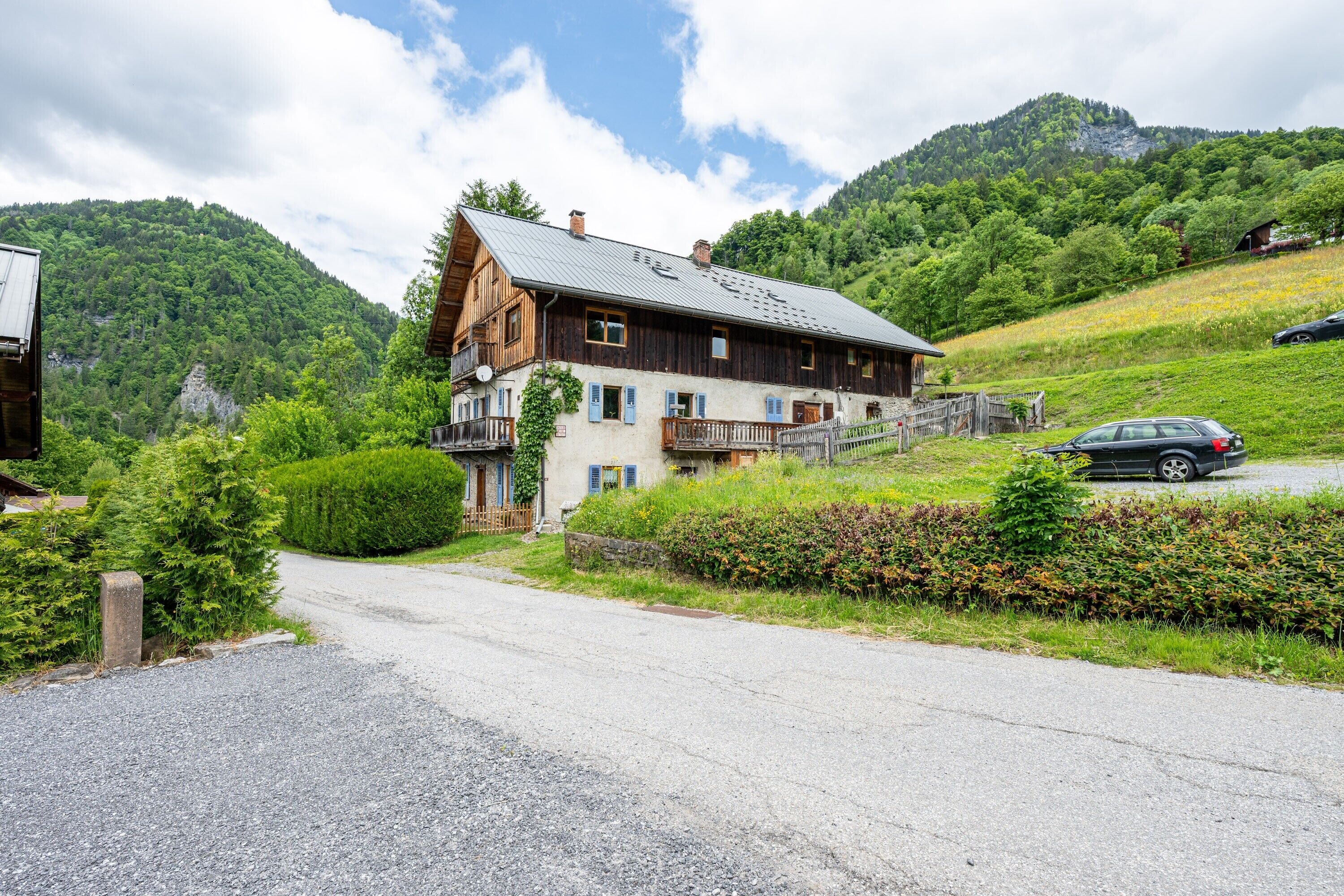 Une maison en bois à l’allure rustique, dotée d’un balcon et entourée d’une végétation luxuriante, avec une chaîne de montagnes en toile de fond.
