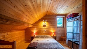 Ein gemütliches Schlafzimmer in einer Blockhütte mit schrägem Dach, einem Bett mit karierter Decke, zwei Nachttischlampen, einem kleinen Fenster und einer integrierten Sitzbank.