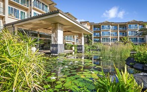 A hotel with a pool surrounded by water lilies and tropical plants, featuring multiple balconies and a covered entrance.