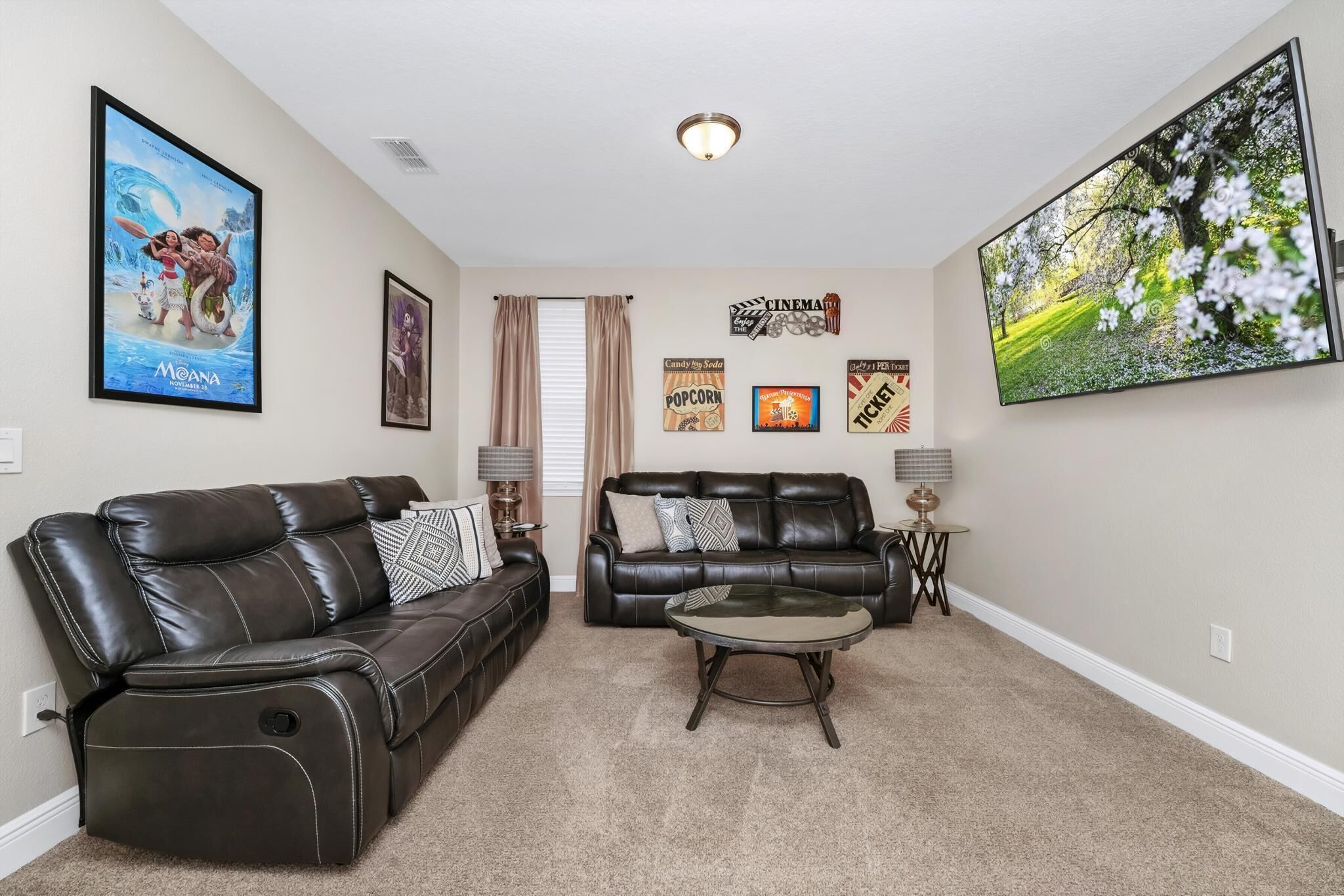 A living room with two black leather sofas, a coffee table, and a large TV screen. There are movie posters on the wall and a window with curtains.