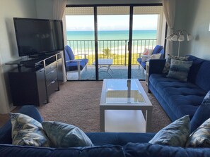 En stue med TV, sofa og sofabord, med udsigt over en strand og hav.
