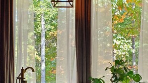 A room with a bathtub, a bench, and a potted plant near a window with sheer curtains.