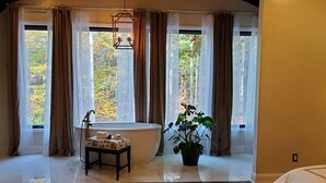 A spacious room with a high ceiling, large windows, a bed, a table with a plant, and a chandelier.